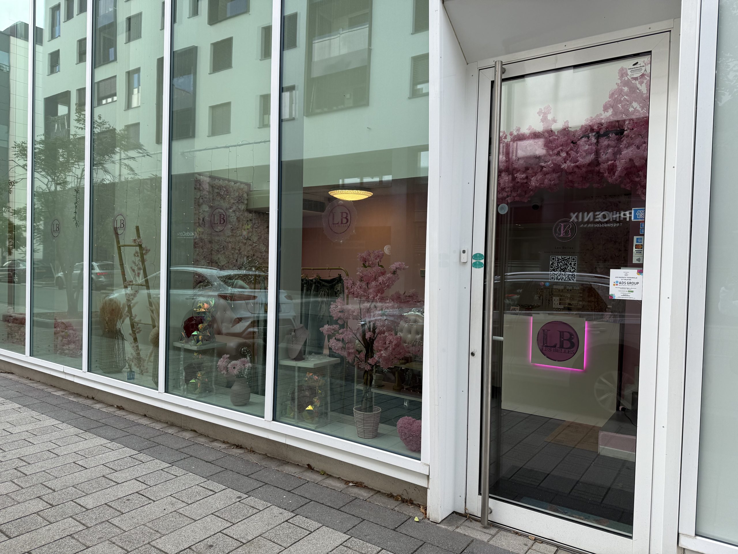 The shopfront of Les Belles in Belval, a beauty salon with a pink neon LB logo on the glass door, card payment and contactless stickers, and a window display of pink artificial cherry blossom, a flower wall, a cream armchair, jewellery on stands and a clothing rail inside.