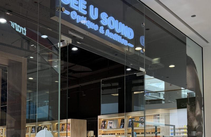 The See U Sound optician in Belval Plaza, with a blue illuminated sign reading Optique & Audition and glasses displayed on wooden shelving inside.