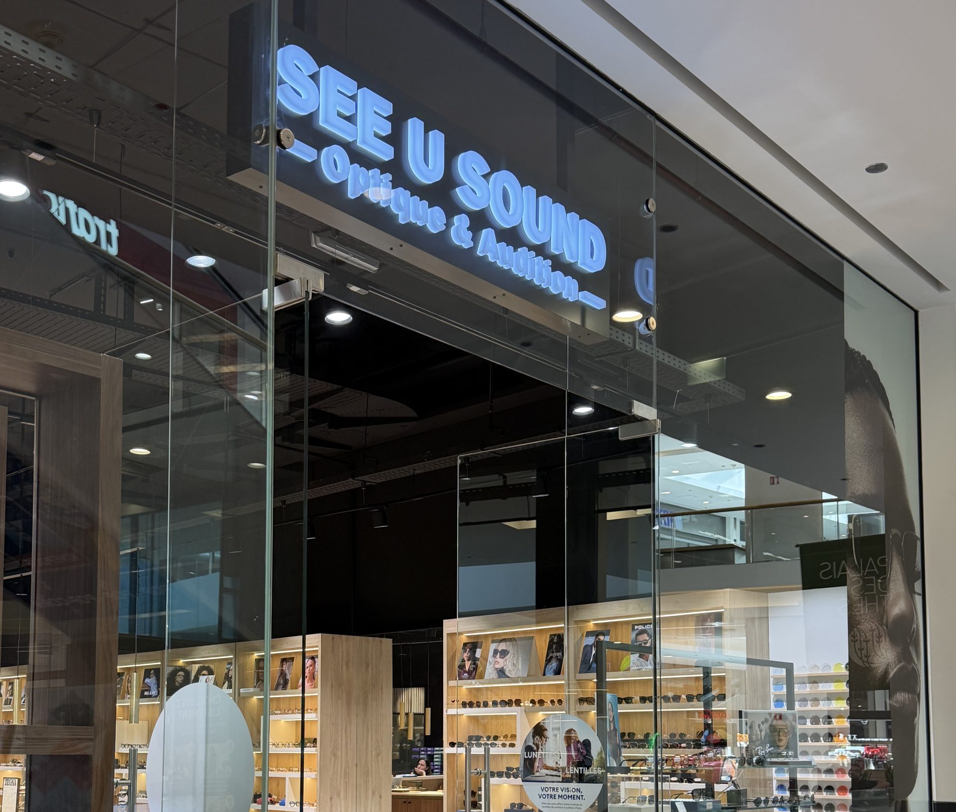 The See U Sound optician in Belval Plaza, with a blue illuminated sign reading Optique & Audition and glasses displayed on wooden shelving inside.