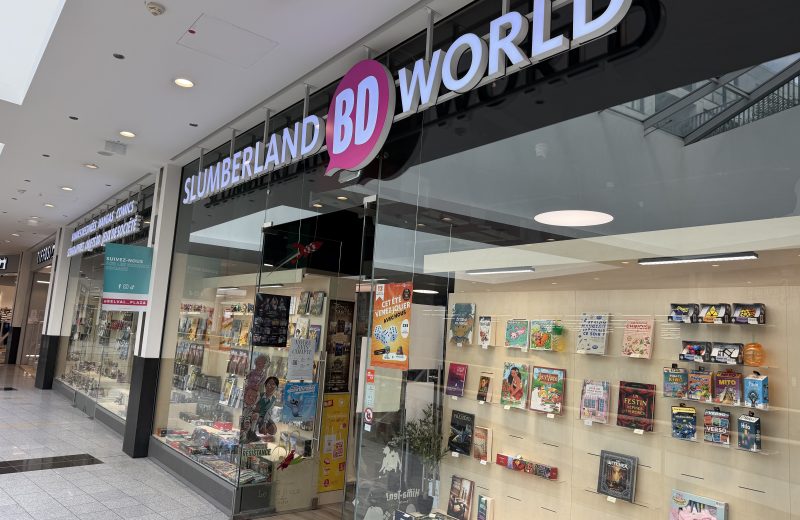 Slumberland BD World shopfront at Belval Plaza, a black and pink storefront with "Slumberland BD World" lettering above a shelf display of comics, manga, board games and collectible figures, a poster advertising a summer games event, and a teal sticker inviting shoppers to follow the centre's own social media accounts.