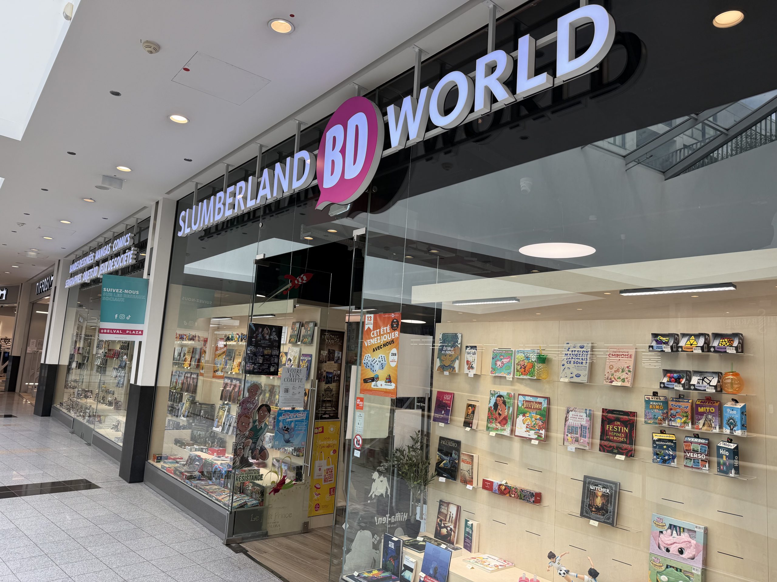 Slumberland BD World shopfront at Belval Plaza, a black and pink storefront with "Slumberland BD World" lettering above a shelf display of comics, manga, board games and collectible figures, a poster advertising a summer games event, and a teal sticker inviting shoppers to follow the centre's own social media accounts.