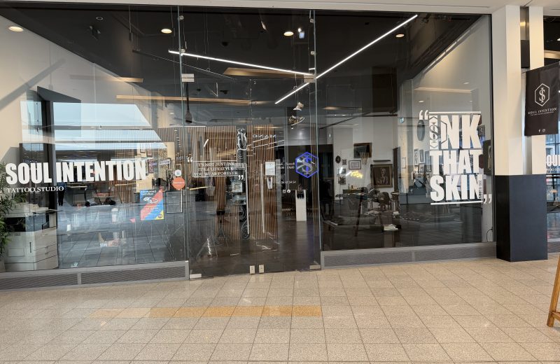Soul Intention Tattoo Studio shopfront at Belval Plaza, "Soul Intention, Tattoo Studio" in large white lettering on the left window, a centre panel with the quote "it's not just ink, it's a lifestyle you wear forever," a Salonkee sticker, a phone number and printed Monday to Saturday opening hours, a right panel with the quote "ink that skin" and "South Ink" worked into the design in smaller type, and a blue neon-style hexagon "S" logo repeated on both the window and a hanging fabric sign further along reading "Soul Intention, Tattoo Studio Luxembourg" with Facebook and Instagram icons.