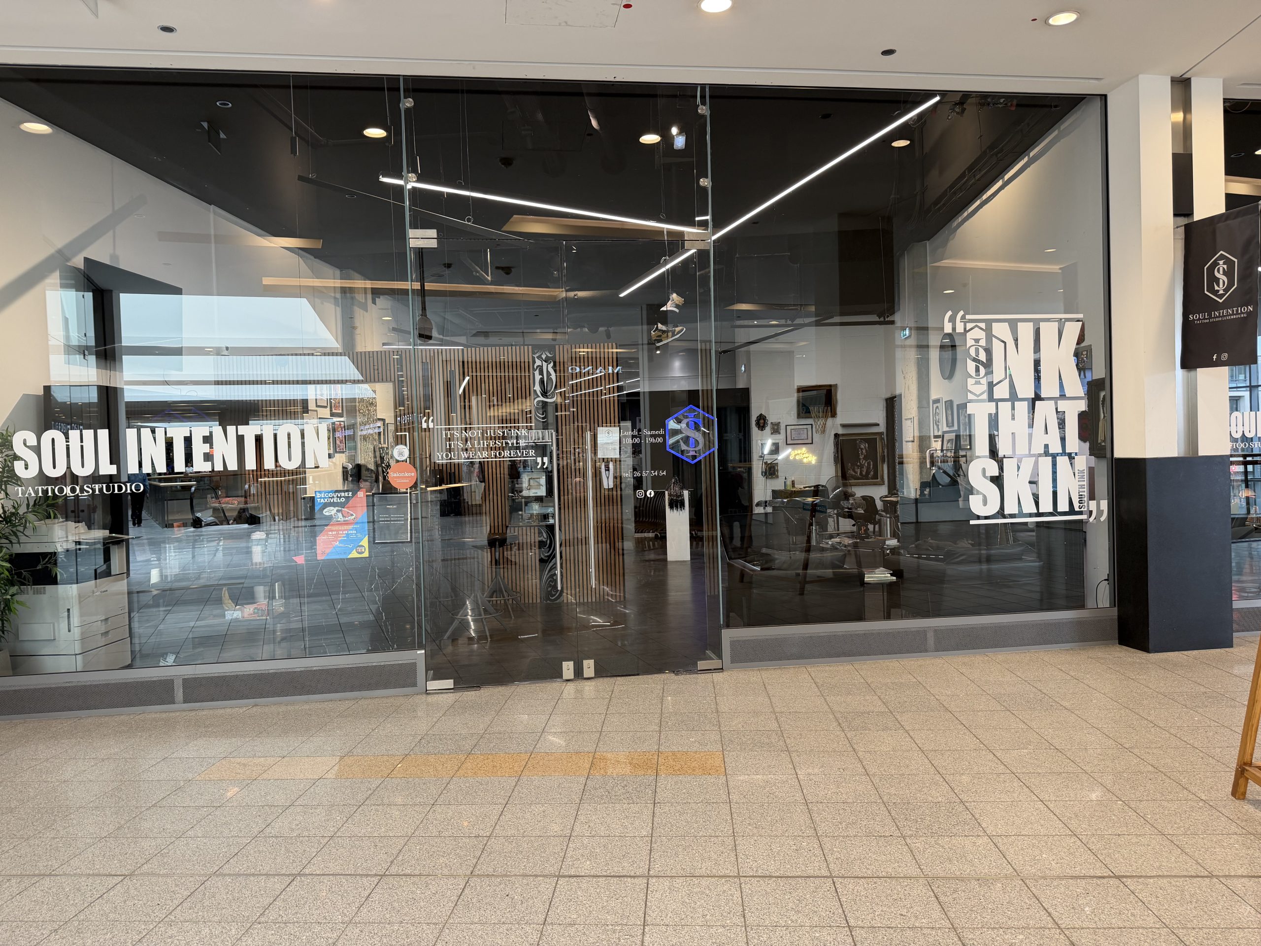 Soul Intention Tattoo Studio shopfront at Belval Plaza, "Soul Intention, Tattoo Studio" in large white lettering on the left window, a centre panel with the quote "it's not just ink, it's a lifestyle you wear forever," a Salonkee sticker, a phone number and printed Monday to Saturday opening hours, a right panel with the quote "ink that skin" and "South Ink" worked into the design in smaller type, and a blue neon-style hexagon "S" logo repeated on both the window and a hanging fabric sign further along reading "Soul Intention, Tattoo Studio Luxembourg" with Facebook and Instagram icons.