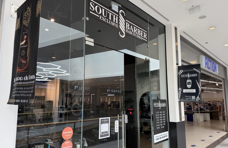 South Barber Exclusive Hair Studios entrance at Belval Plaza, black glass frontage with white lettering reading "South Barber Exclusive Hair Studios" above a scissors and razor strop logo, a black banner reading "Lance de Vous" with a mobile number for a bespoke shave service, a door decal listing hours from Monday to Saturday 10h to 19h with Sunday closed, and a Salonkee five-star "2025 Top Rated" sticker.
