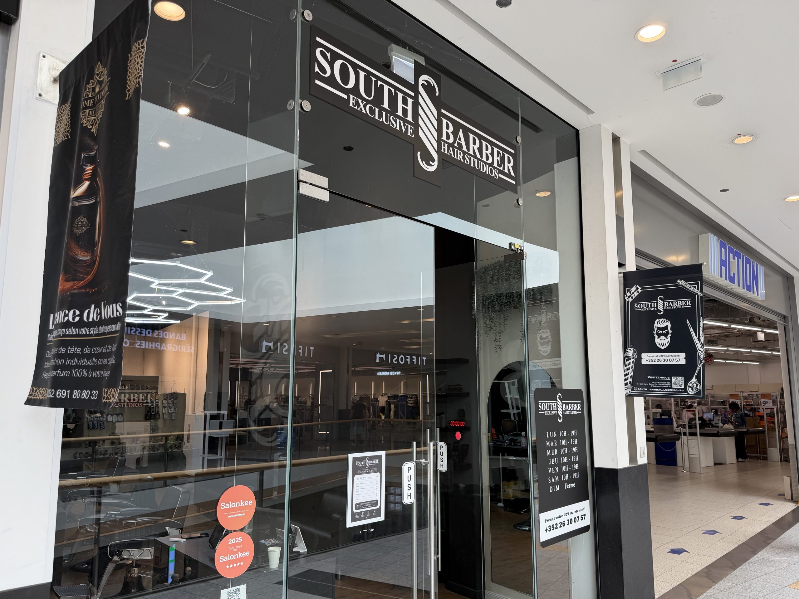 South Barber Exclusive Hair Studios entrance at Belval Plaza, black glass frontage with white lettering reading "South Barber Exclusive Hair Studios" above a scissors and razor strop logo, a black banner reading "Lance de Vous" with a mobile number for a bespoke shave service, a door decal listing hours from Monday to Saturday 10h to 19h with Sunday closed, and a Salonkee five-star "2025 Top Rated" sticker.