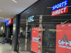 The Sports Direct and USC shopfront at Belval Plaza, with illuminated signs, brand logos and red sale posters in the window.