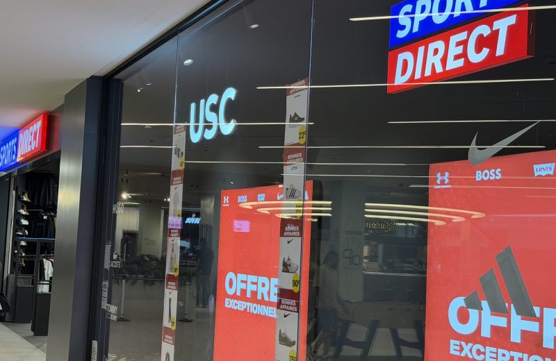 The Sports Direct and USC shopfront at Belval Plaza, with illuminated signs, brand logos and red sale posters in the window.