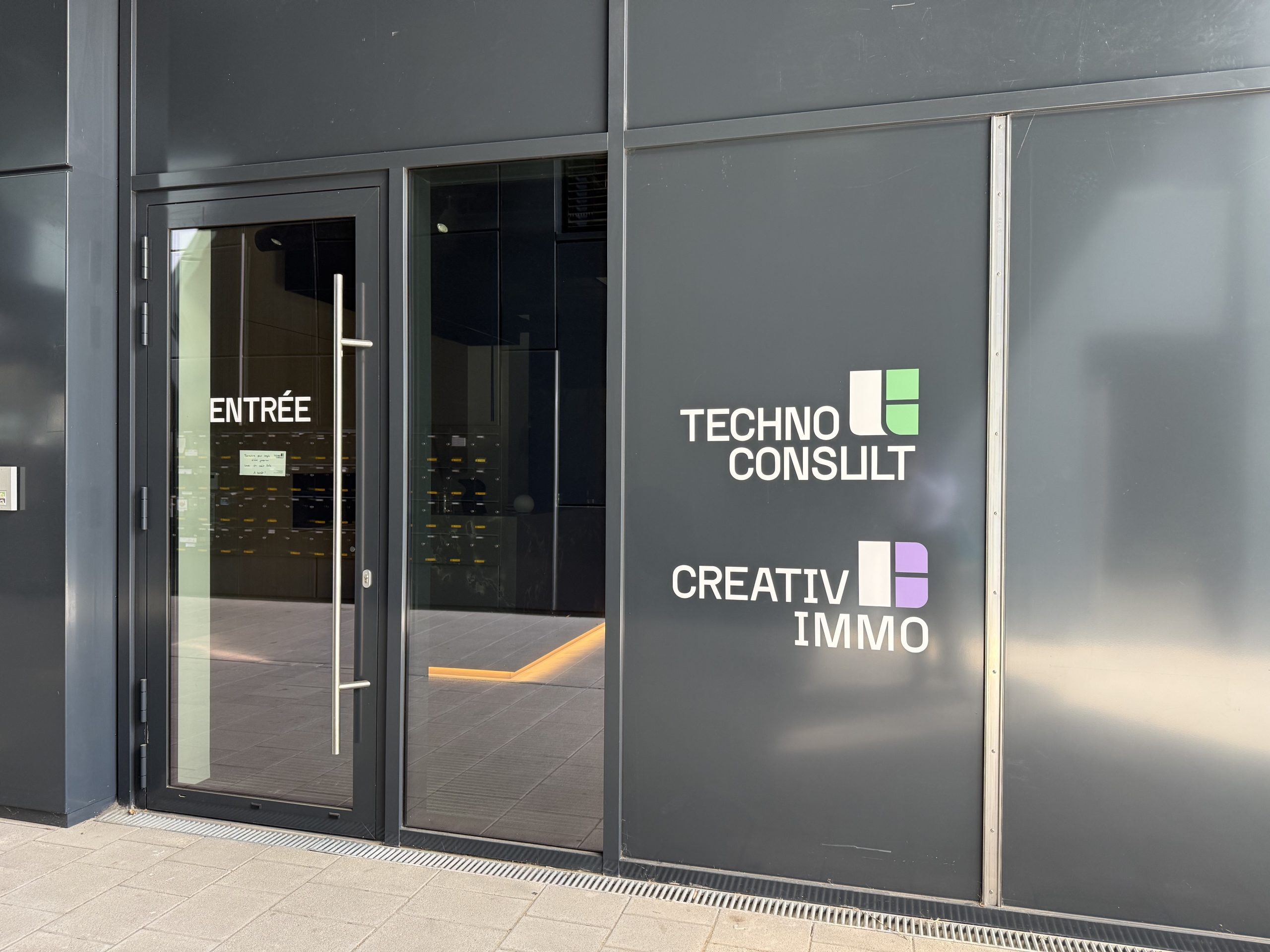 The shared entrance to Résidence Gateway on boulevard du Jazz, a grey panelled frontage with a glass door marked ENTRÉE opening onto a wall of letterboxes, and the Techno Consult and Creativ Immo logos applied to the panel beside it.