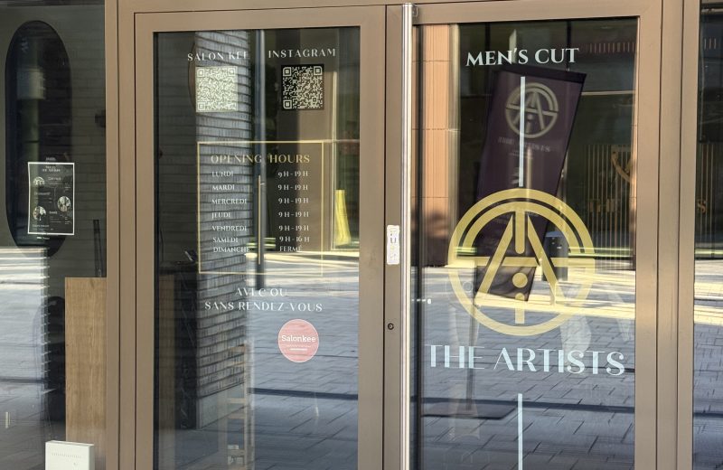 The Artists Barber salon doors, with a gold circular emblem and "Men's Cut" and "The Artists" lettering, opening hours listed in gold text for Monday to Saturday with Sunday marked closed, and QR codes for Salonkee booking and Instagram, with an interior poster listing haircut prices including a students category visible through the glass.