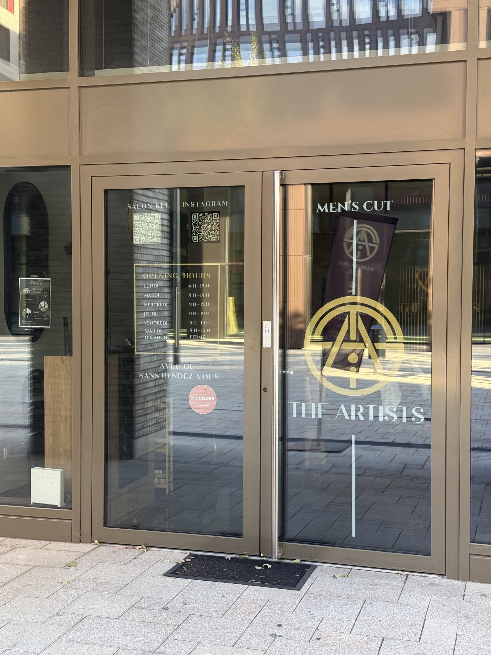 The Artists Barber salon doors, with a gold circular emblem and "Men's Cut" and "The Artists" lettering, opening hours listed in gold text for Monday to Saturday with Sunday marked closed, and QR codes for Salonkee booking and Instagram, with an interior poster listing haircut prices including a students category visible through the glass.