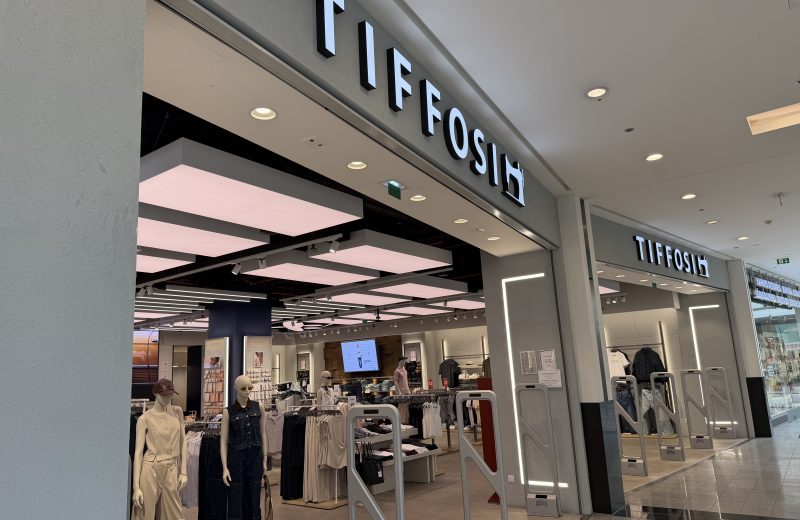 Tiffosi clothing shop entrance at Belval Plaza, grey and white storefront with backlit "Tiffosi" lettering, mannequins dressed in denim and casualwear near the entrance, and security gates either side of the door.