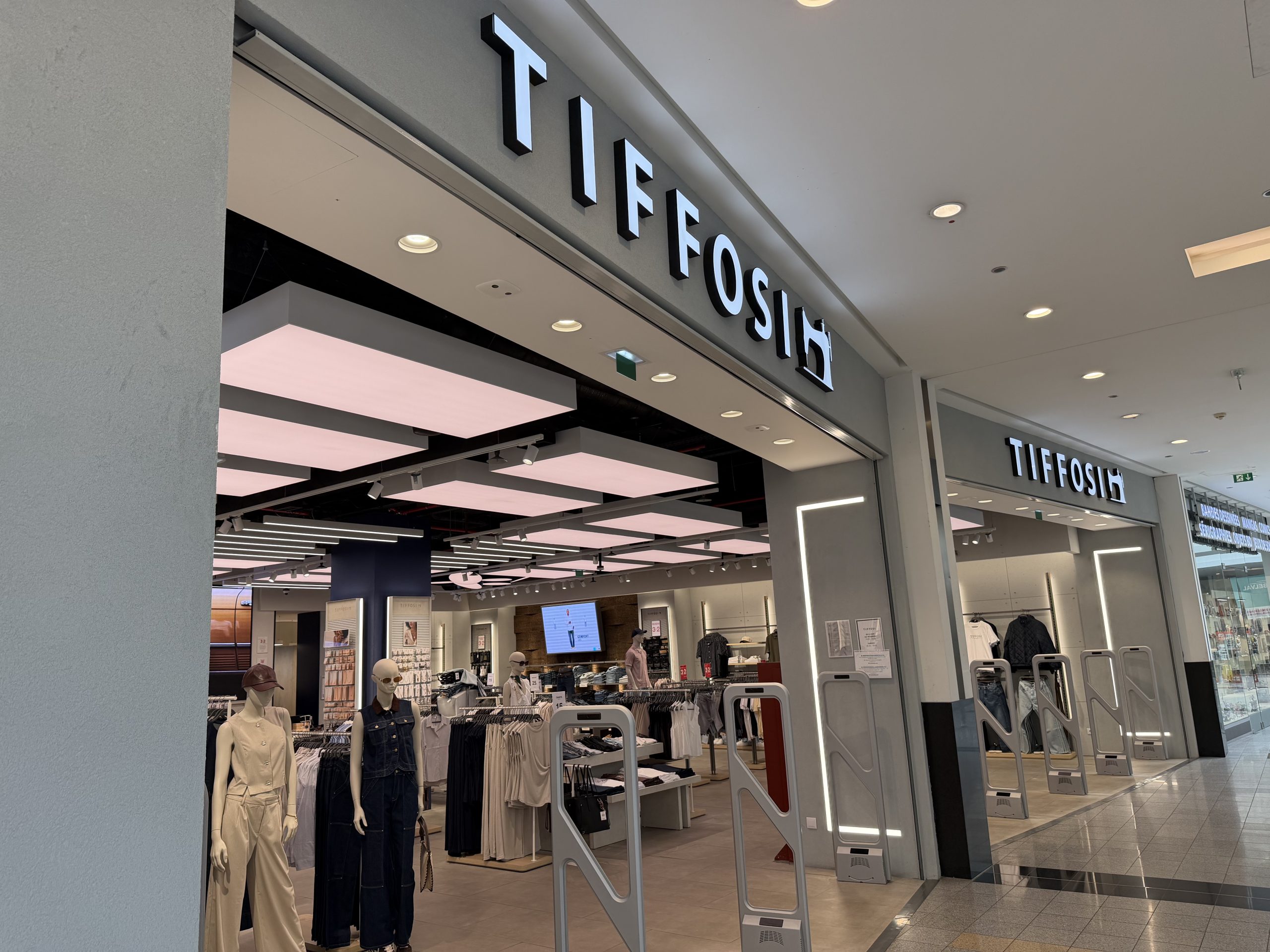 Tiffosi clothing shop entrance at Belval Plaza, grey and white storefront with backlit "Tiffosi" lettering, mannequins dressed in denim and casualwear near the entrance, and security gates either side of the door.