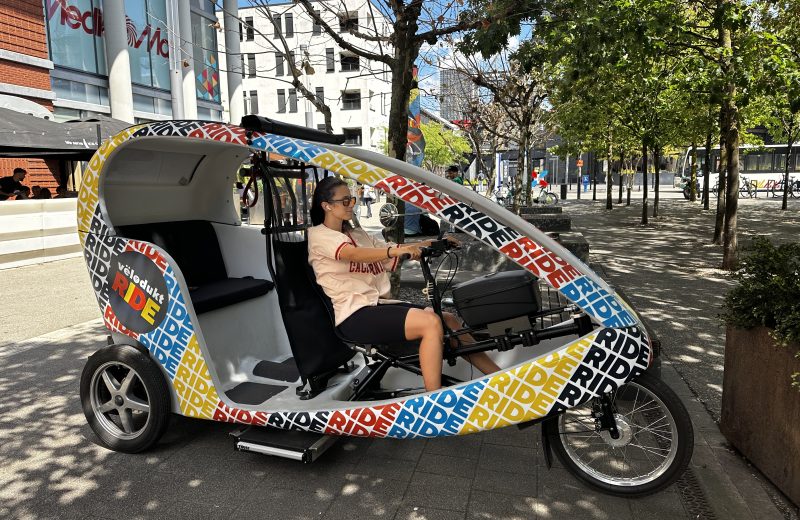 The Vëlodukt Ride bike taxi parked outside MediaMarkt in Belval, its driver seated at the handlebars