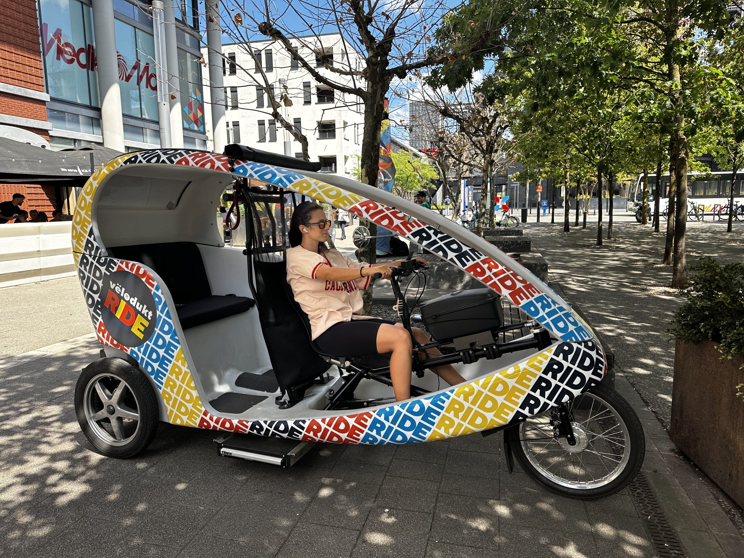The Vëlodukt Ride bike taxi parked outside MediaMarkt in Belval, its driver seated at the handlebars