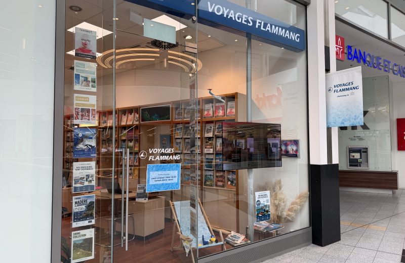 The Voyages Flammang shopfront at Belval Plaza, with the Flammang wordmark above the entrance and a printed poster in the window giving opening hours for Monday to Friday and Saturday.