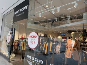 White House clothing shop entrance at Belval Plaza, black fascia reading "White House, The Fashion Store," mannequins in autumn coats and blazers near the entrance, and a red "Promo jusqu'à -50%" sticker on the glass.
