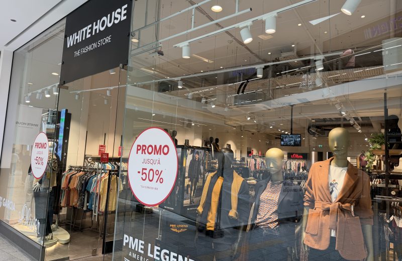 White House clothing shop entrance at Belval Plaza, black fascia reading "White House, The Fashion Store," mannequins in autumn coats and blazers near the entrance, and a red "Promo jusqu'à -50%" sticker on the glass.