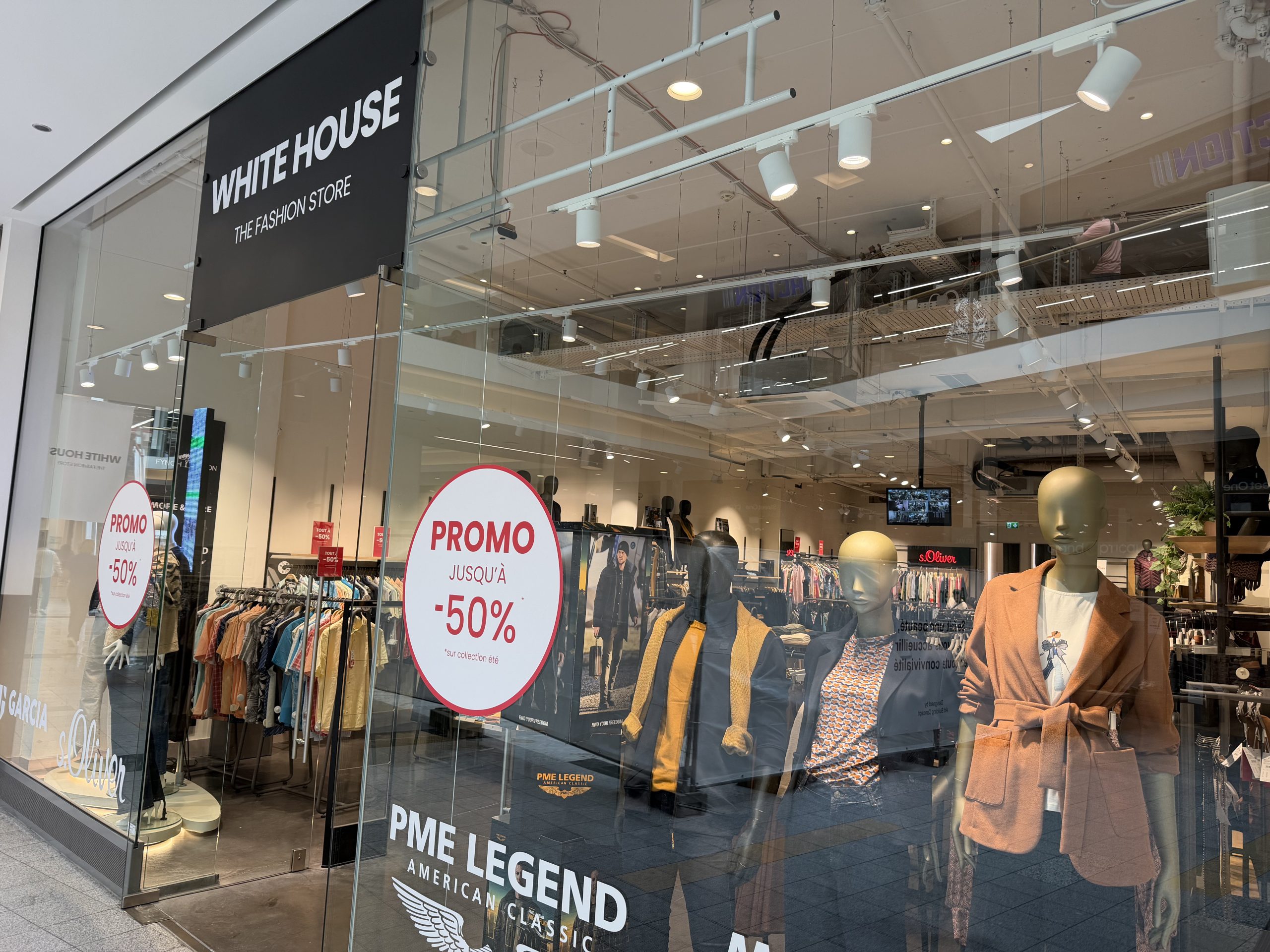 White House clothing shop entrance at Belval Plaza, black fascia reading "White House, The Fashion Store," mannequins in autumn coats and blazers near the entrance, and a red "Promo jusqu'à -50%" sticker on the glass.