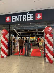 The entrance to Delhaize Belval on reopening morning, with a red carpet, red and white balloon columns either side and shoppers walking in under the Entrée sign