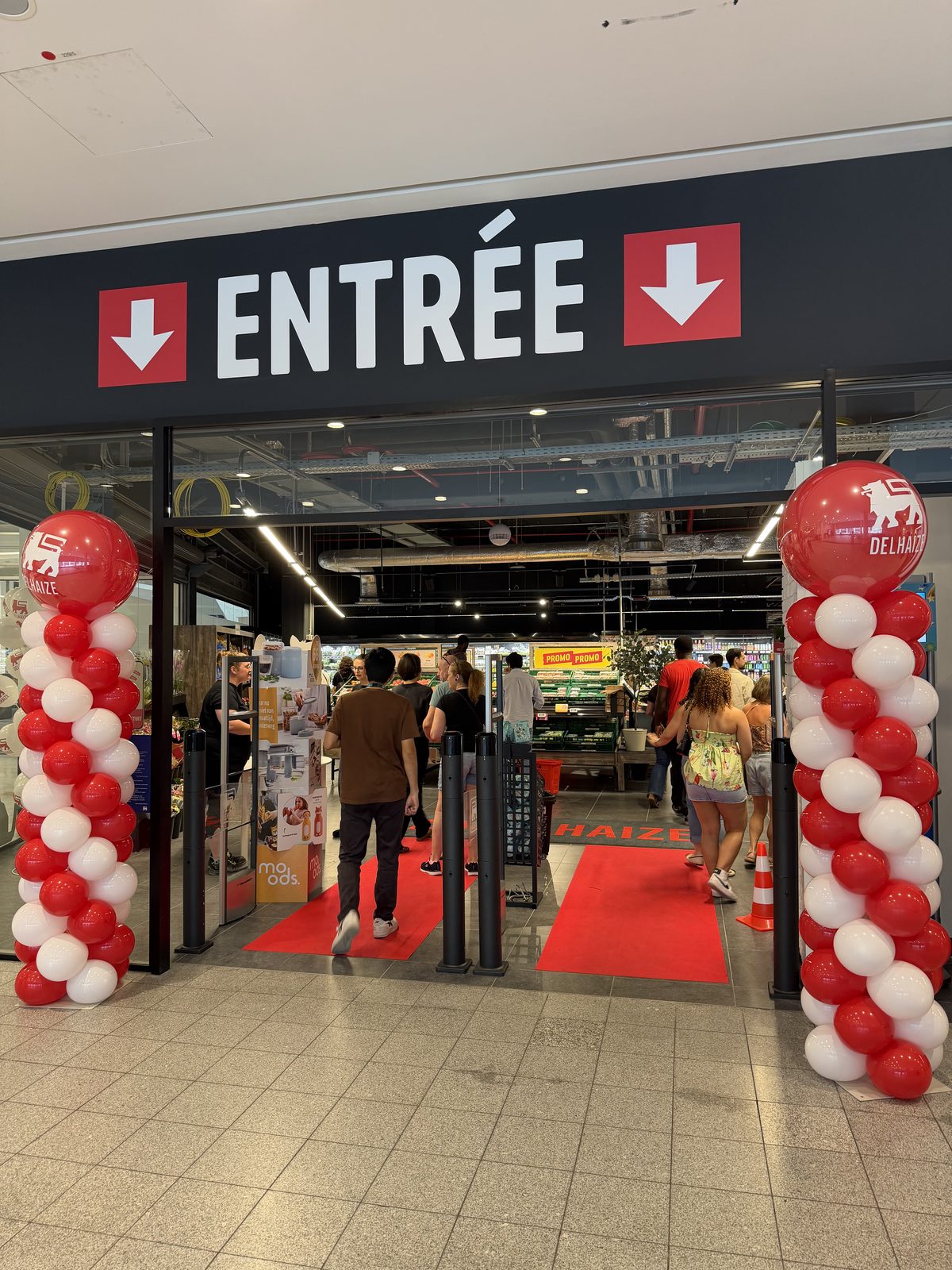The entrance to Delhaize Belval on reopening morning, with a red carpet, red and white balloon columns either side and shoppers walking in under the Entrée sign