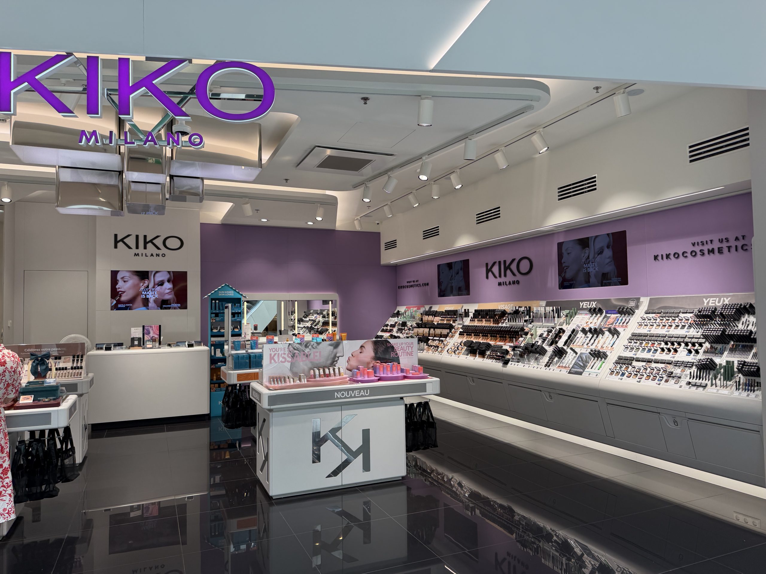 The Kiko Milano shopfront in Belval Plaza, with the purple KIKO MILANO sign above the entrance and rows of makeup testers lining the wall inside, including a table of new lipstick launches.