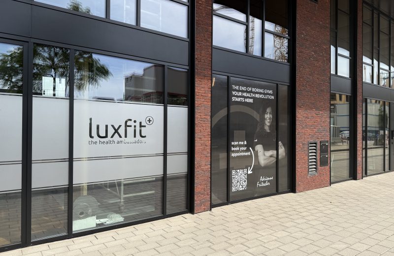 The Luxfit shopfront in Belval, a black-framed glass frontage between red brick piers, with the lowercase luxfit logo and the words "the health ambassadors" on frosted window film, and a dark panel alongside reading "the end of boring gyms, your health revolution starts here" with a QR code for booking.