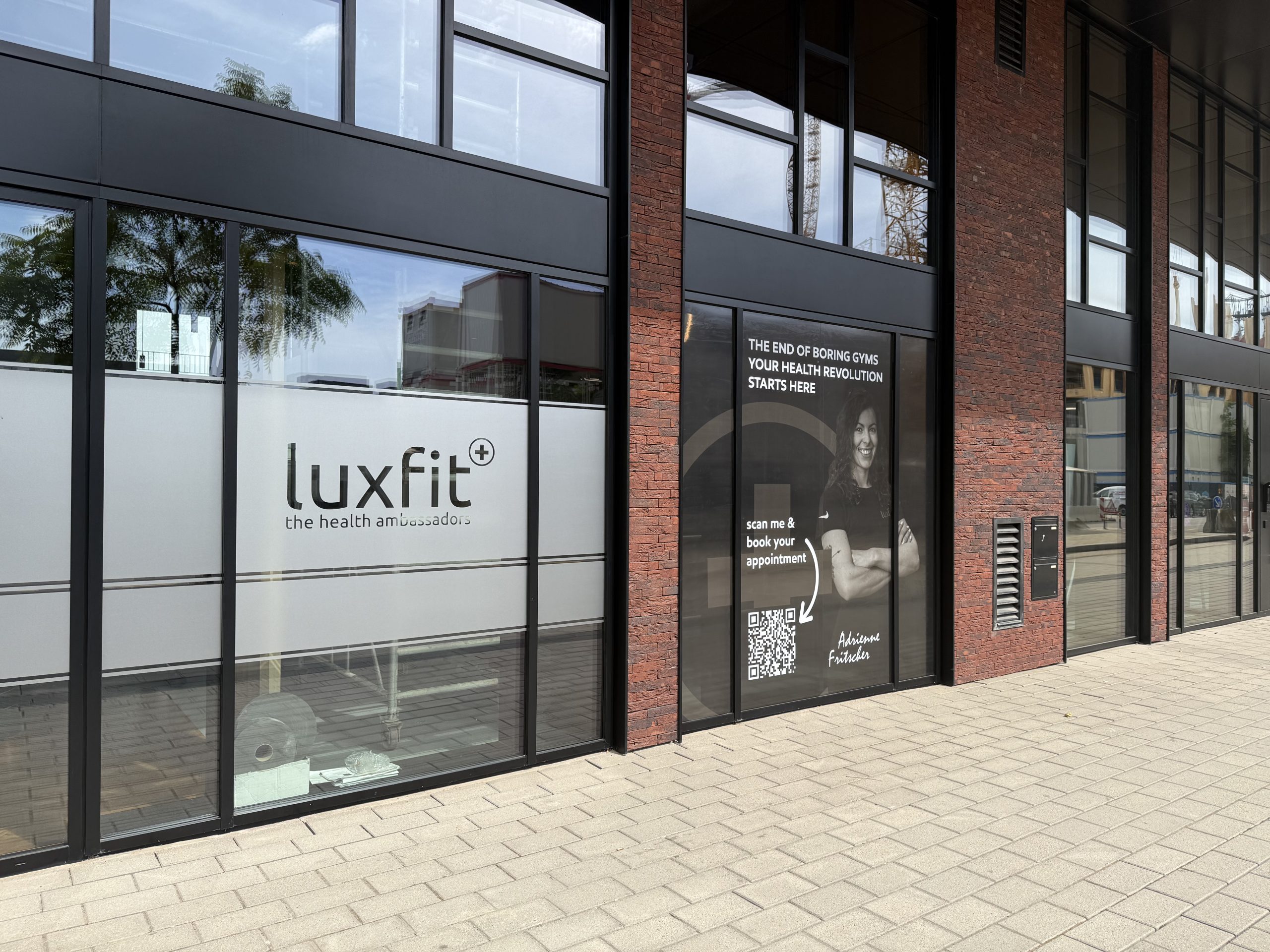 The Luxfit shopfront in Belval, a black-framed glass frontage between red brick piers, with the lowercase luxfit logo and the words "the health ambassadors" on frosted window film, and a dark panel alongside reading "the end of boring gyms, your health revolution starts here" with a QR code for booking.
