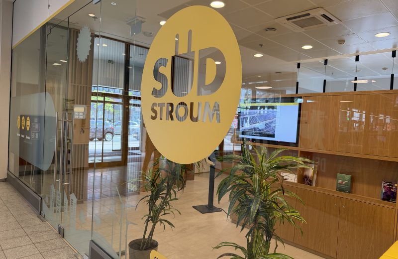 The Sudstroum counter inside Belval Plaza, seen through the glass frontage, with a large yellow circular logo reading SUD STROUM with a plug motif, a wood-panelled interior with a screen and brochure shelves, and pictograms on the door for electricity, internet and television.