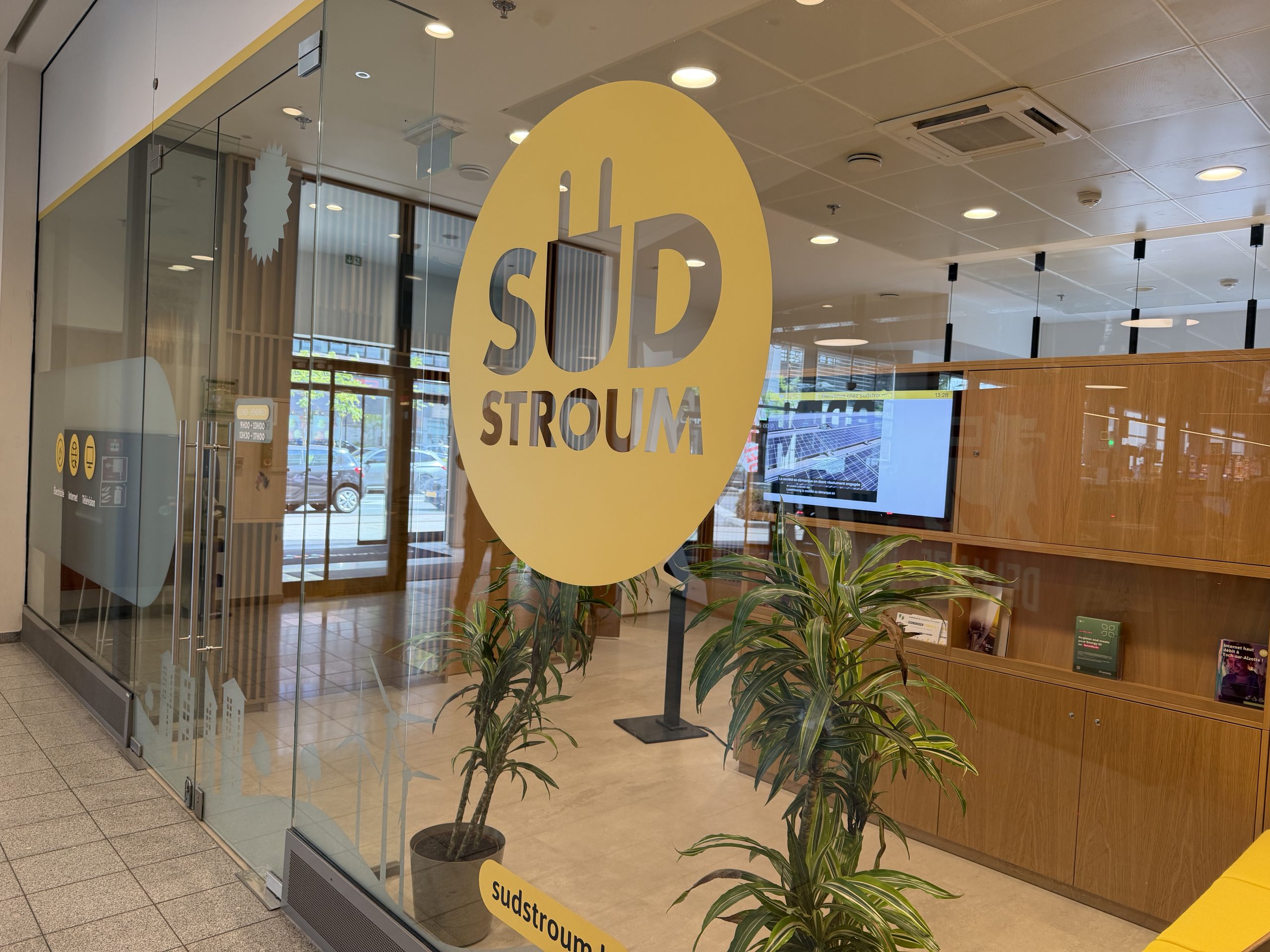 The Sudstroum counter inside Belval Plaza, seen through the glass frontage, with a large yellow circular logo reading SUD STROUM with a plug motif, a wood-panelled interior with a screen and brochure shelves, and pictograms on the door for electricity, internet and television.