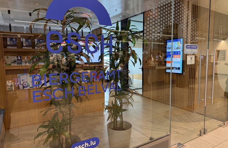 The Biergeramt Esch Belval citizens' office inside Belval Plaza, seen through its glass frontage, with the blue esch.lu logo and BIERGERAMT ESCH BELVAL lettering on the glass, an opening hours decal reading Lundi-Vendredi 9H00-17H00, brochure shelves and an information screen behind.
