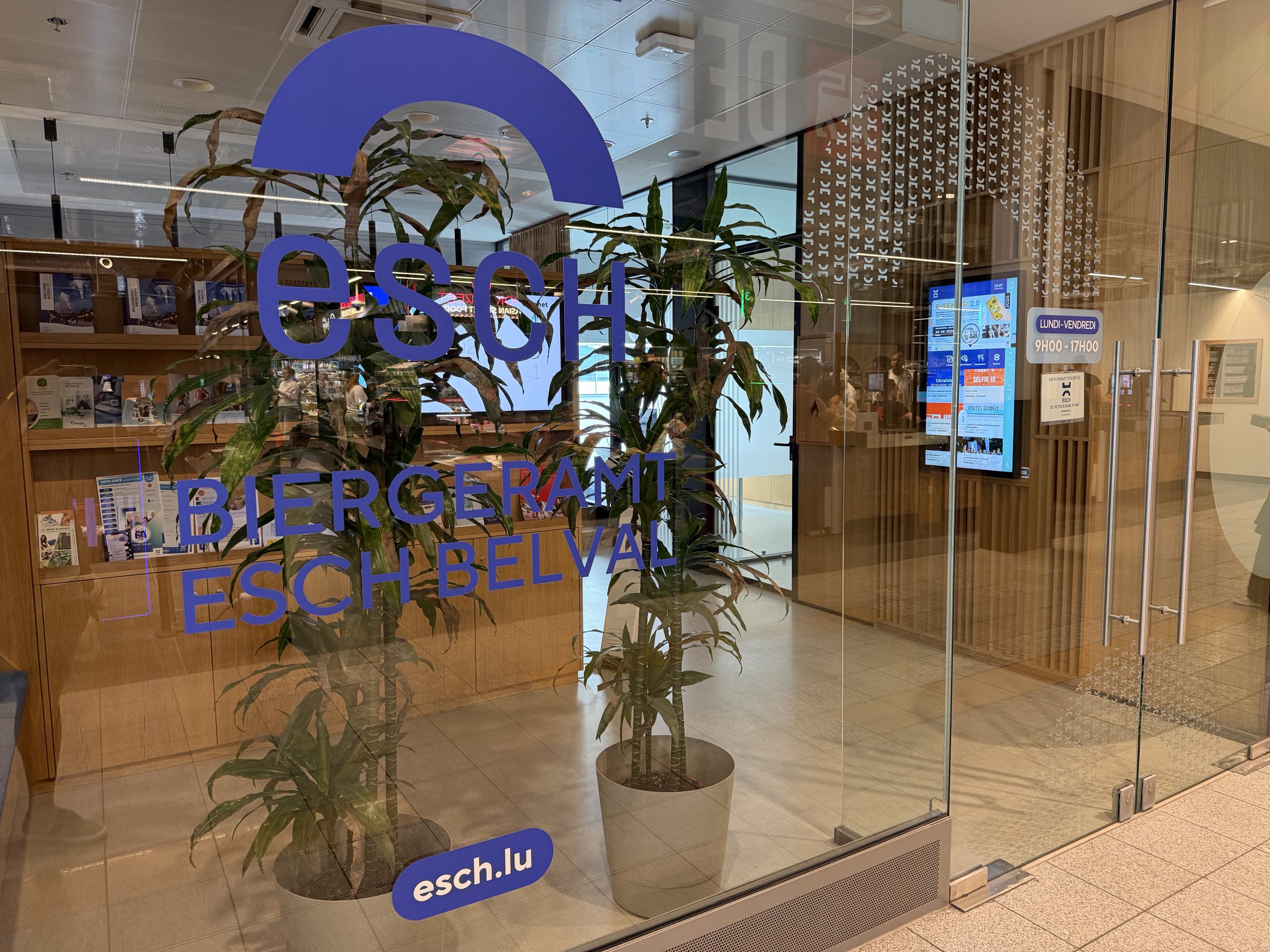 The Biergeramt Esch Belval citizens' office inside Belval Plaza, seen through its glass frontage, with the blue esch.lu logo and BIERGERAMT ESCH BELVAL lettering on the glass, an opening hours decal reading Lundi-Vendredi 9H00-17H00, brochure shelves and an information screen behind.