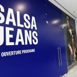 The Salsa Jeans hoarding inside Belval Plaza I: a deep blue wall with SALSA JEANS in large white letters and OUVERTURE PROCHAINE beneath, a denim campaign photo of two models on the right, and a Belval Plaza sign at the corridor corner.