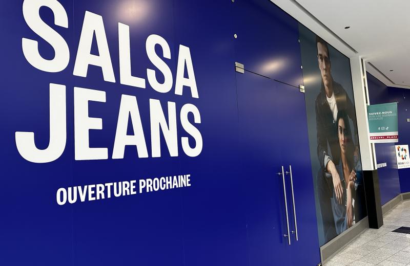The Salsa Jeans hoarding inside Belval Plaza I: a deep blue wall with SALSA JEANS in large white letters and OUVERTURE PROCHAINE beneath, a denim campaign photo of two models on the right, and a Belval Plaza sign at the corridor corner.