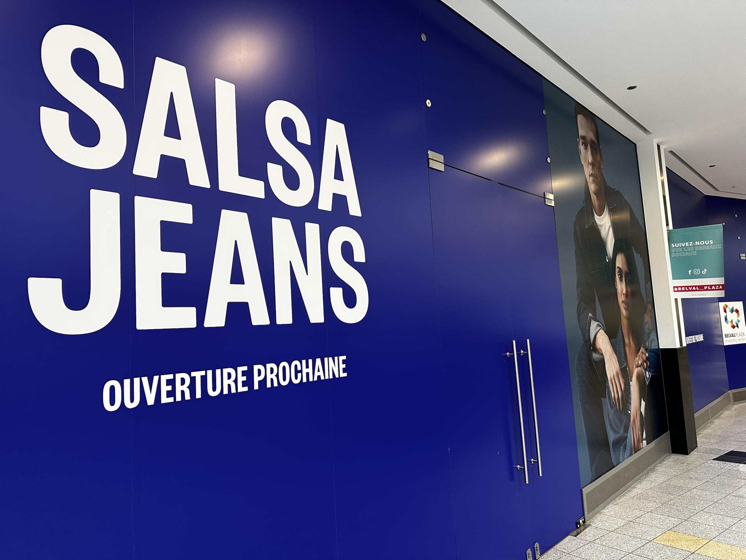 The Salsa Jeans hoarding inside Belval Plaza I: a deep blue wall with SALSA JEANS in large white letters and OUVERTURE PROCHAINE beneath, a denim campaign photo of two models on the right, and a Belval Plaza sign at the corridor corner.