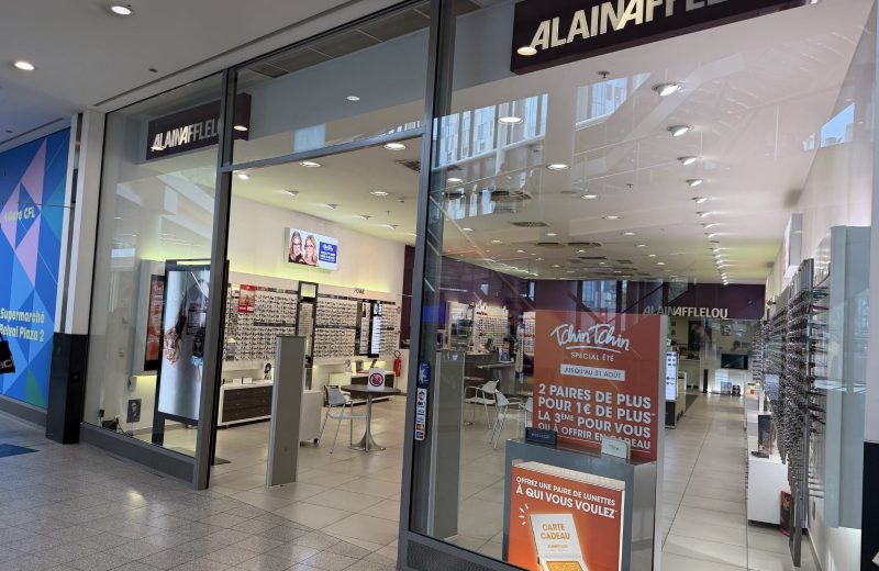 The Alain Afflelou shopfront at Belval Plaza (shown mirrored in the photograph, shot through glass), with the Afflelou wordmark above the entrance and a blue promotional wall panel reading "Belval Plaza 2" visible behind the display.