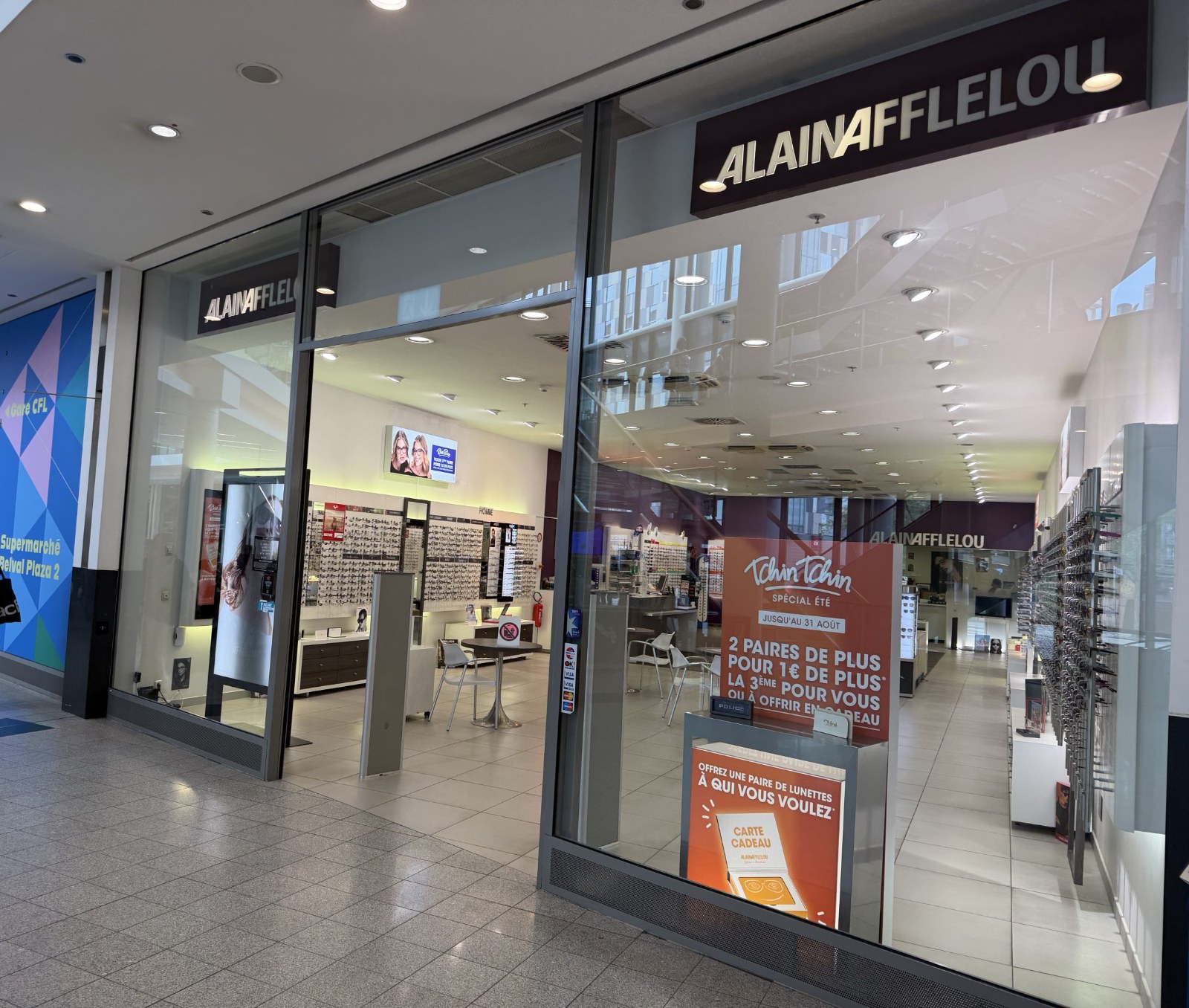 The Alain Afflelou shopfront at Belval Plaza (shown mirrored in the photograph, shot through glass), with the Afflelou wordmark above the entrance and a blue promotional wall panel reading "Belval Plaza 2" visible behind the display.