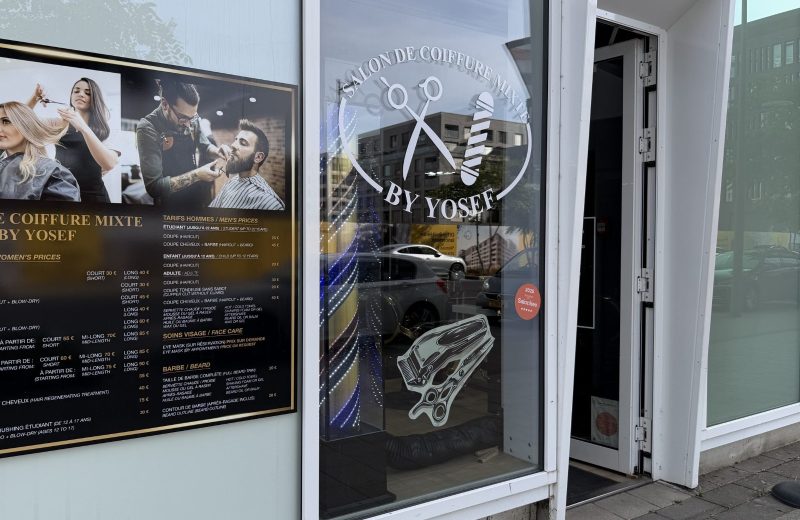 The By Yosef salon in Belval, with a large price board in French and English in the window and the salon logo etched on the glass.