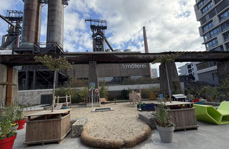 The Esch Beeval square in Belval: raised wooden planters and flower pots around a sand playground, under the old conveyor bridge, with the Möllerei and the blast furnaces behind.