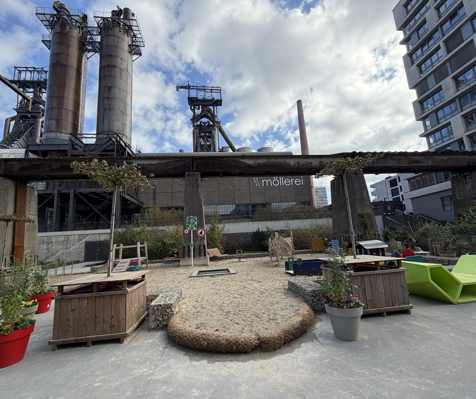 The Esch Beeval square in Belval: raised wooden planters and flower pots around a sand playground, under the old conveyor bridge, with the Möllerei and the blast furnaces behind.