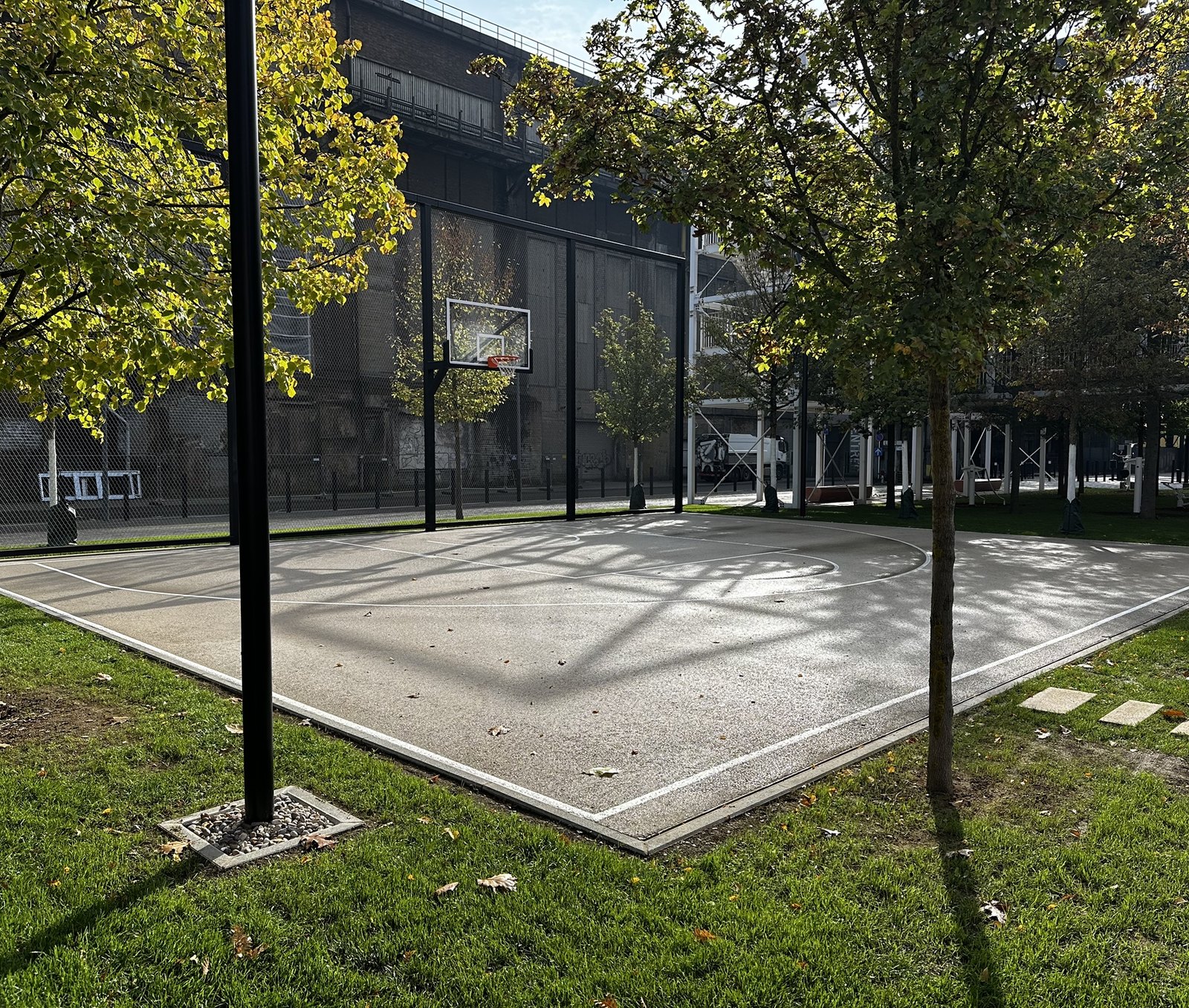 The half basketball court at Belval Outdoor, fenced and ringed with young trees, with the dark steel of the blast furnace works behind it.