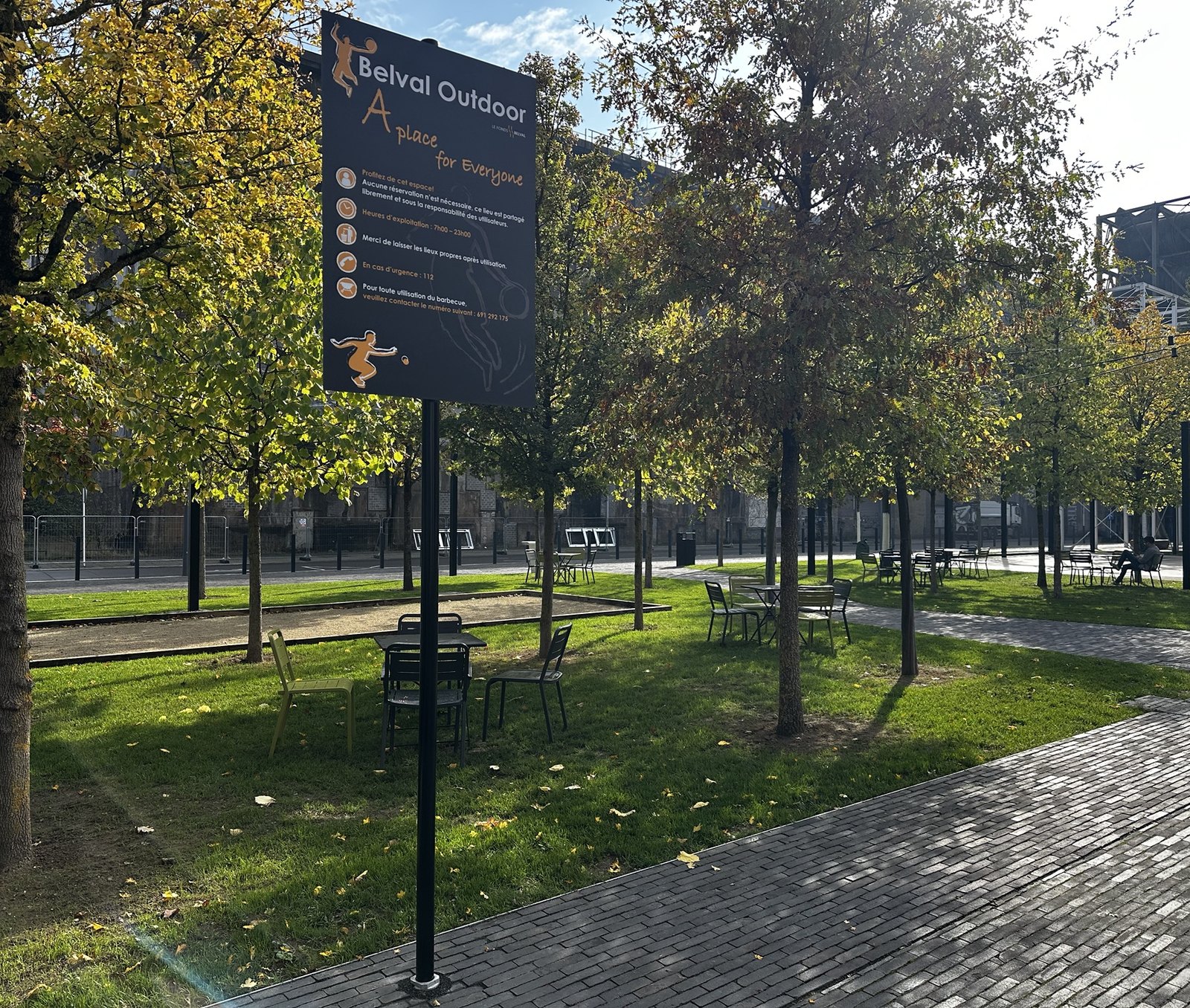 La pelouse de Belval Outdoor, tables et chaises sous de jeunes arbres, la piste de pétanque derriére et le panneau d’information au premier plan.
