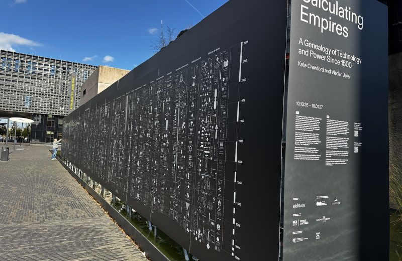 Calculating Empires at Socle C in Belval: the title panel and the long black timeline wall in the sun, with a visitor reading it and the Restaurant de l’Université behind