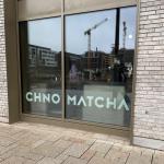 The Chno Matcha window in Belvaux, the name in pale green on the glass, with a coffee machine, cups and jugs on the counter inside and cranes reflected in the pane.