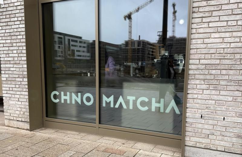 The Chno Matcha window in Belvaux, the name in pale green on the glass, with a coffee machine, cups and jugs on the counter inside and cranes reflected in the pane.