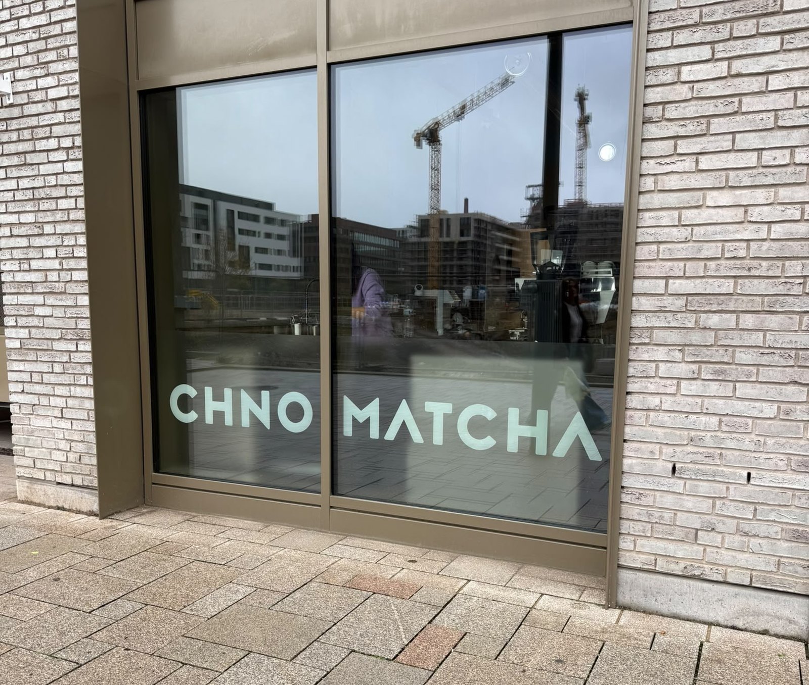 The Chno Matcha window in Belvaux, the name in pale green on the glass, with a coffee machine, cups and jugs on the counter inside and cranes reflected in the pane.
