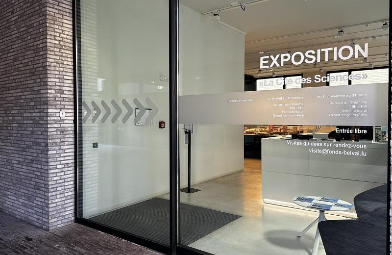 The entrance to the Cité des Sciences exhibition in Belval, EXPOSITION lettering above the glass, the reception desk inside and the seasonal opening hours printed on the window.