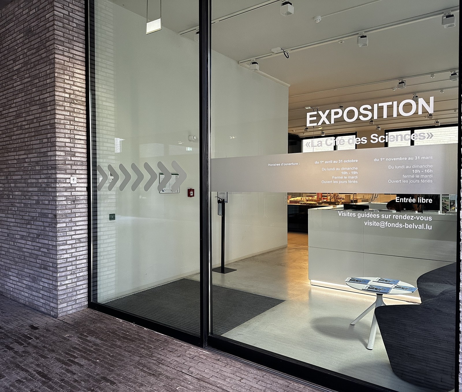 The entrance to the Cité des Sciences exhibition in Belval, EXPOSITION lettering above the glass, the reception desk inside and the seasonal opening hours printed on the window.