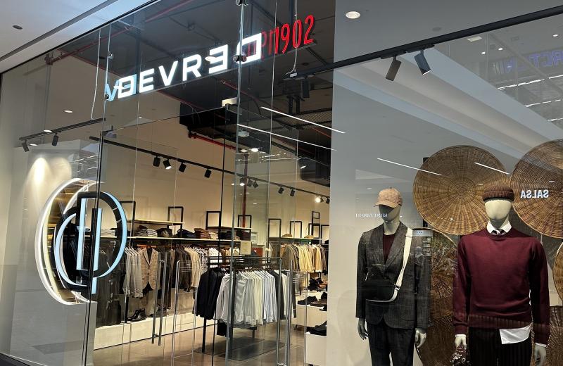 The Devred 1902 shopfront in Belval Plaza, the illuminated DEVRED 1902 sign above the glass, the monogram on the window and rails of menswear inside.