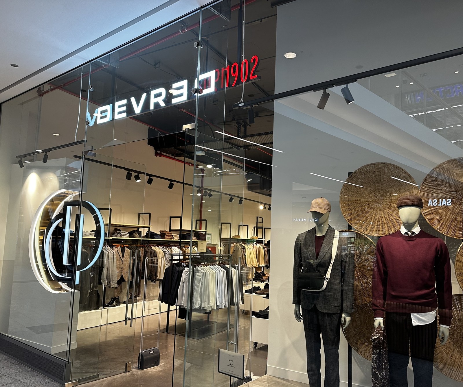 The Devred 1902 shopfront in Belval Plaza, the illuminated DEVRED 1902 sign above the glass, the monogram on the window and rails of menswear inside.