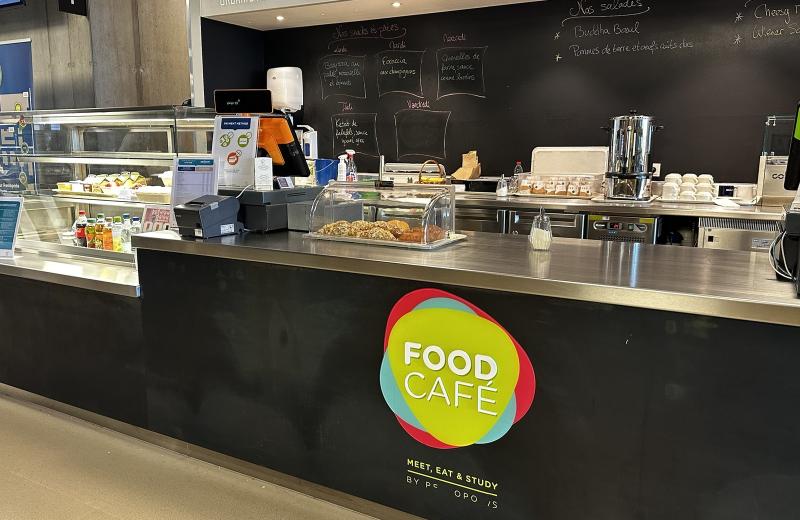 The Food Café counter on the Belval campus, its yellow and red roundel on the front, pastries and drinks in the chiller and a chalkboard of the week’s snacks behind.